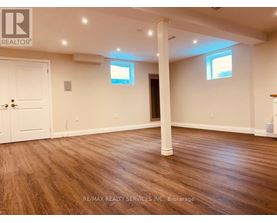 A room with wooden flooring and white walls, with a door on the left and a window on the right.