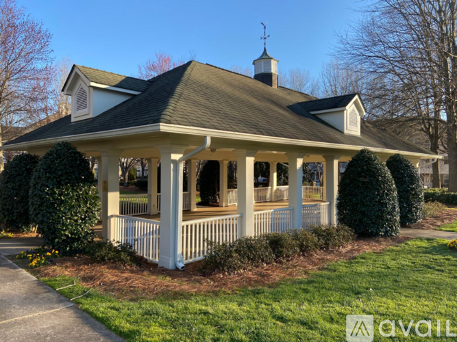 A white gazebo with a black roof and a weather vane on top is surrounded by a white picket fence and green bushes.