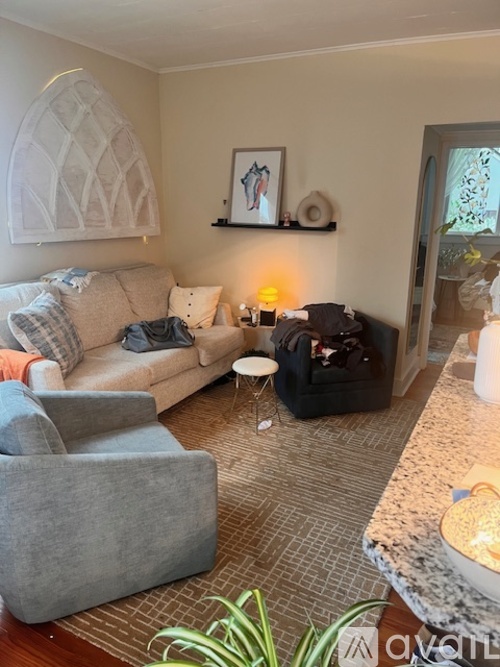 A living room with a grey couch, a grey armchair, a black ottoman, and a white table with a lamp on it.