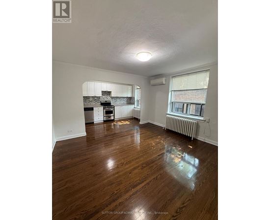 A room with wooden floors and a kitchen area in the background.
