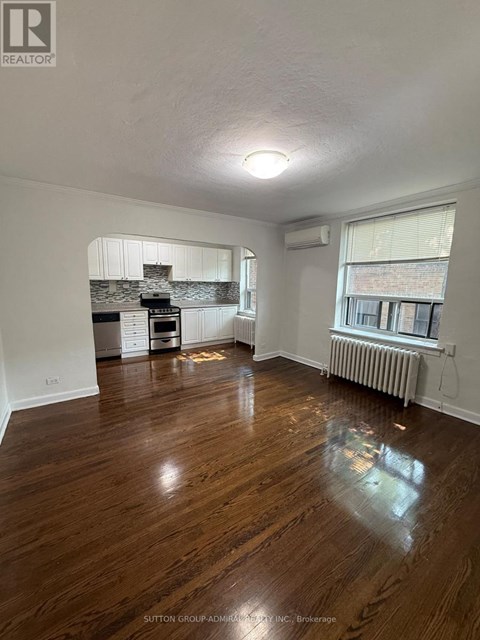 A room with wooden floors and a kitchen area in the background.