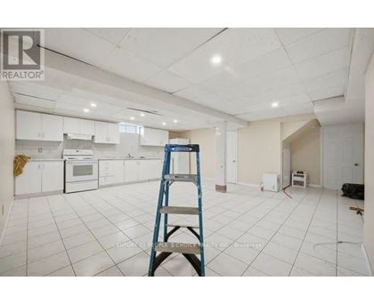 A room with white tiled floors and walls, a ladder, and a kitchen area with white cabinets and appliances.