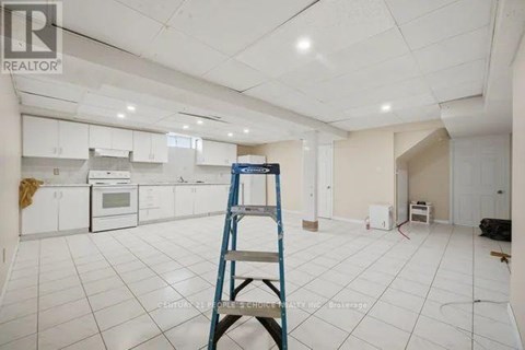 A room with white tiled floors and walls, a ladder, and a kitchen area with white cabinets and appliances.