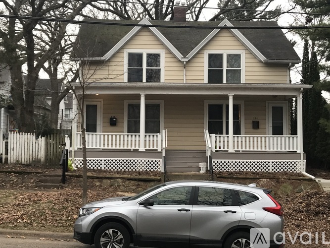 A silver car is parked in front of a two-story house with a white porch.