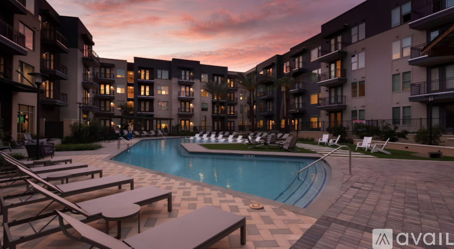 A pool surrounded by lounge chairs and apartment buildings at dusk.