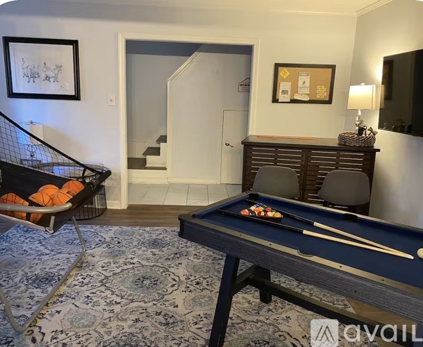 A pool table with a blue cloth and a white cue ball is in the foreground of a room with a television, a chair, and a staircase in the background.