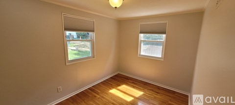 A room with two windows and wooden floors is available for rent.