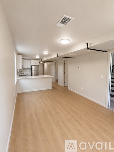 A room with wooden floors and white walls, with a kitchen area visible in the background.