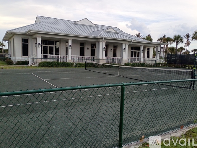 A tennis court is surrounded by a green fence and a white building with a grey roof.