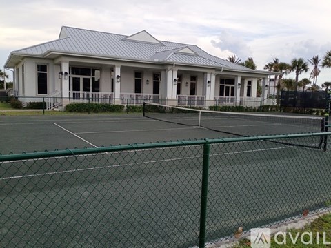 A tennis court is surrounded by a green fence and a white building with a grey roof.