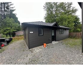 A small house with a black door and windows is surrounded by a gravel driveway.