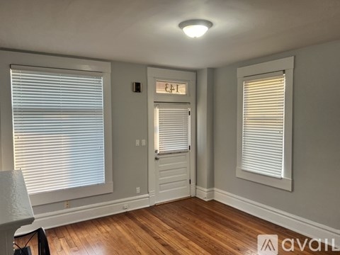 A room with wooden floors and white walls, featuring a window with blinds and a door with a window above it.