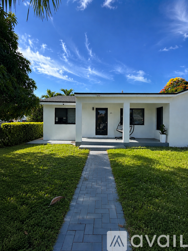 A white house with a black door and windows is surrounded by greenery.