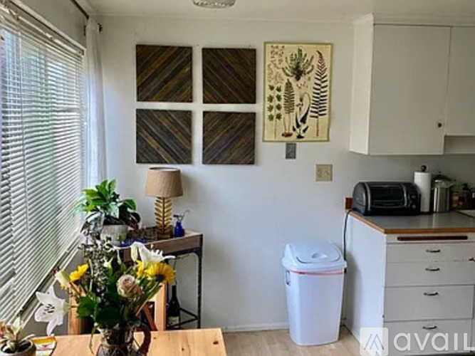 A white trash can sits in a kitchen next to a table with a vase of flowers on it.