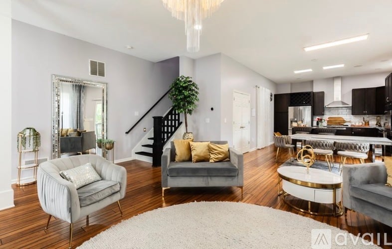 A modern living room with a grey sofa, a grey armchair, a grey coffee table, a black staircase, a black kitchen, a white rug, a chandelier, and a mirror.