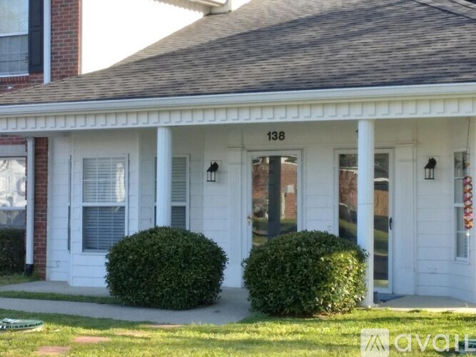 A white house with a porch and two bushes in front.