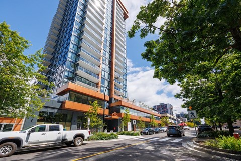 A modern building with a glass facade is surrounded by trees and vehicles on the street.