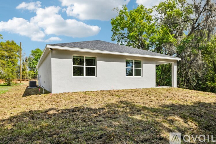 A small white house with a grey roof and a black chimney is situated on a grassy area with trees in the background.