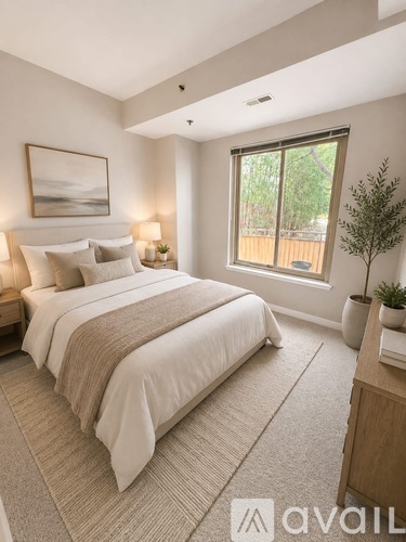 A bedroom with a large bed, a small table with a plant, and a window showing a wooden fence outside.