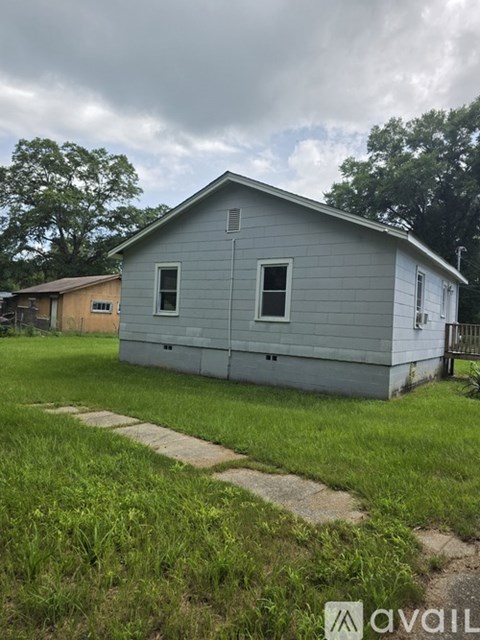 A grey house with a white door and windows is surrounded by green grass and a cloudy sky.