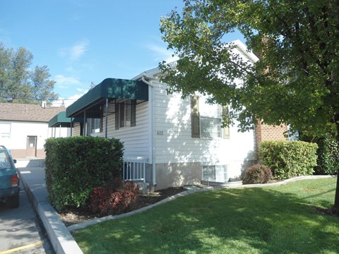 A white house with a green awning is surrounded by a green lawn and trees.