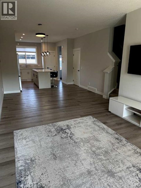 A large, open living room with a grey rug in the foreground and a kitchen area in the background.