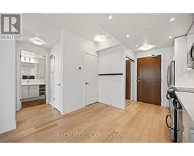 A spacious room with wooden floors and white walls, with a kitchen visible in the background.