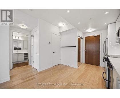 A spacious room with wooden floors and white walls, with a kitchen visible in the background.