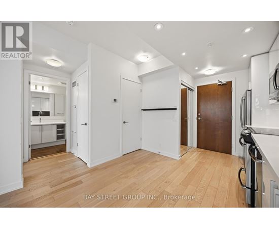 A spacious room with wooden floors and white walls, with a kitchen visible in the background.