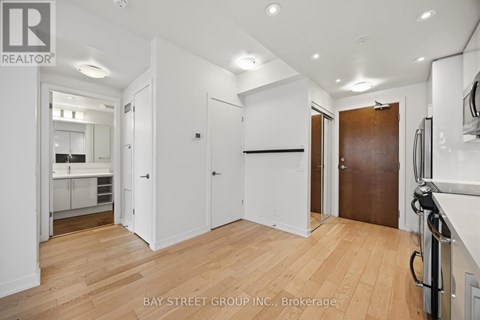 A spacious room with wooden floors and white walls, with a kitchen visible in the background.