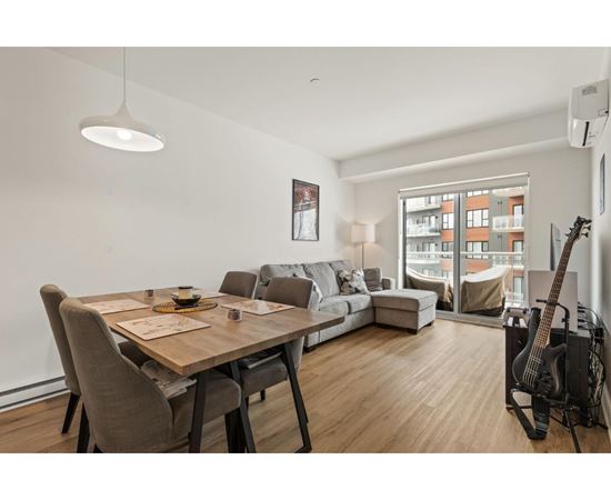 A modern living room with a wooden dining table and chairs, a couch, a guitar, and a window overlooking a building.