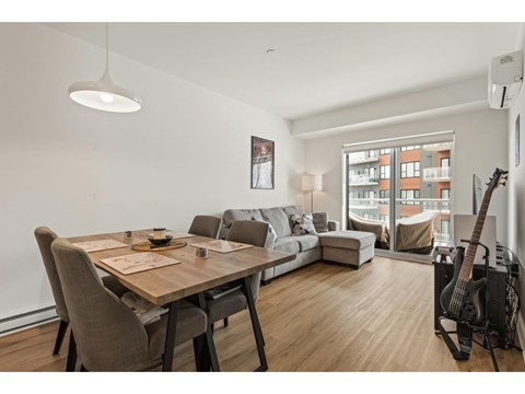 A modern living room with a wooden dining table and chairs, a couch, a guitar, and a window overlooking a building.