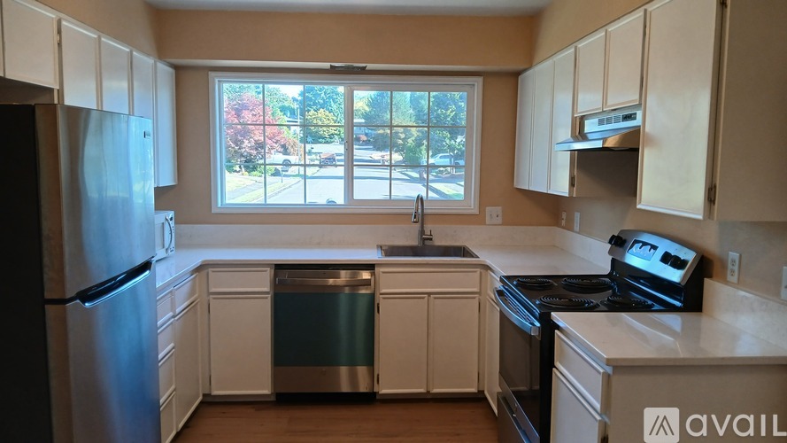 A kitchen with white cabinets and appliances, a window with a view of trees, and a wooden floor.
