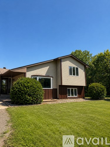 A house with a brown and beige exterior is surrounded by green grass and trees.