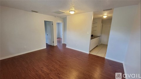 A room with wooden flooring and white walls, with a door on the left and a kitchen area on the right.