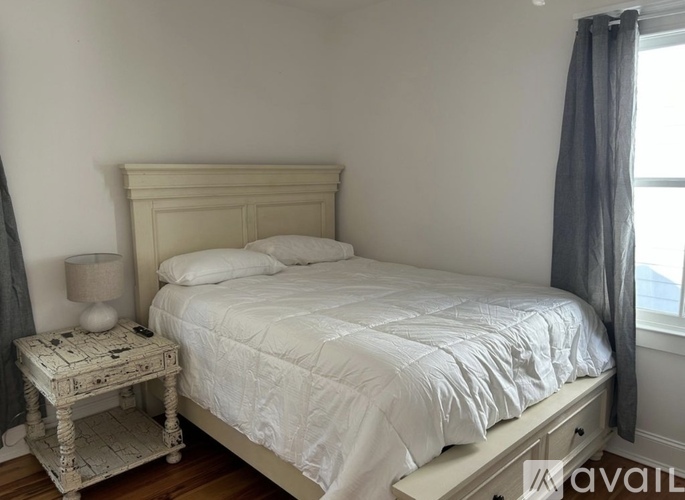 A white bed with a white bedspread and pillow is in a room with a window and a grey curtain.