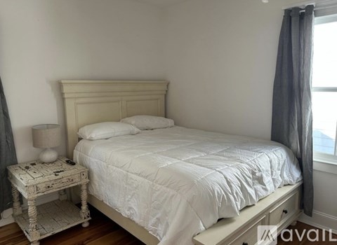 A white bed with a white bedspread and pillow is in a room with a window and a grey curtain.