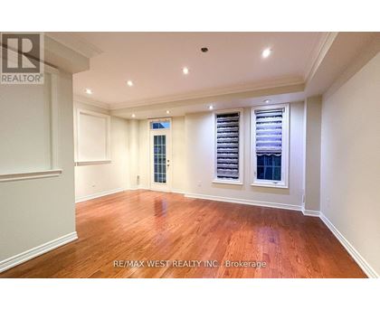 A spacious room with wooden floors and white walls, featuring a large window with blinds and a smaller window with a view of a building.