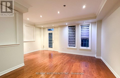 A spacious room with wooden floors and white walls, featuring a large window with blinds and a smaller window with a view of a building.