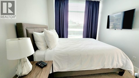 A bedroom with a bed, nightstand, lamp, and a flat screen TV mounted on the wall.