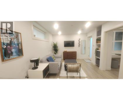 A living room with a grey sofa, a black chair, a white side table, a wooden cabinet, a TV, a plant, and a framed picture on the wall.