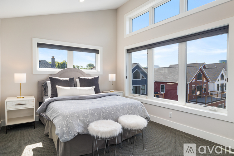 A bedroom with a large bed, a window with a view of houses, and a nightstand with a lamp.