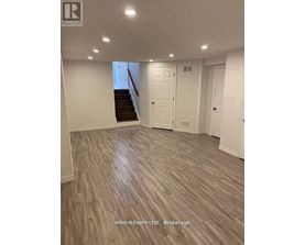 A spacious room with wood flooring and a staircase in the background.