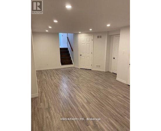 A spacious room with wood flooring and a staircase in the background.