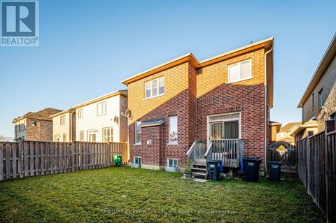 A house with a brown brick exterior and a white window is surrounded by a wooden fence and has a green trash can in the yard.