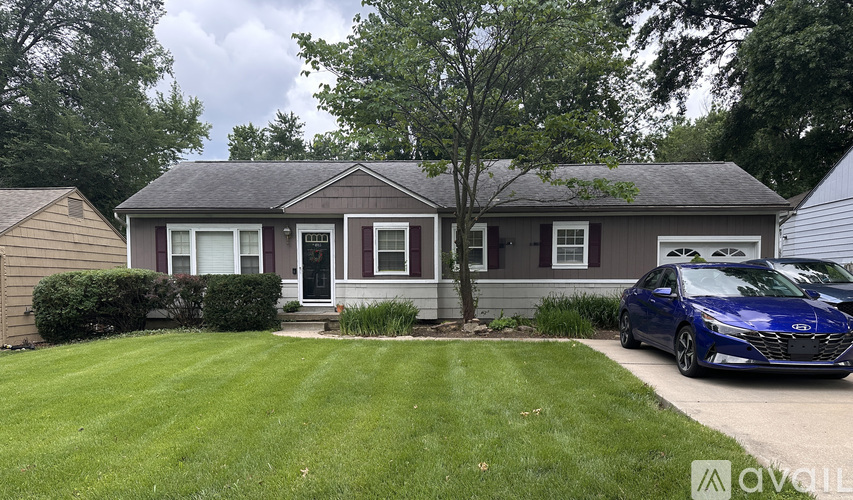 A blue car is parked in front of a house with a tree in the front yard.