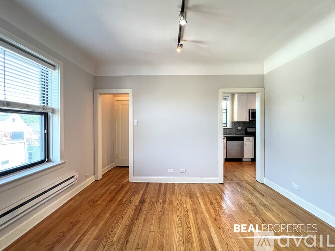A spacious room with wooden flooring and white walls, with a window on the left side.