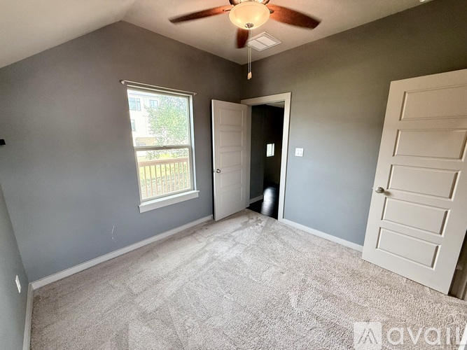 A room with a carpeted floor, a ceiling fan, and a window with a view of a fence and trees outside.