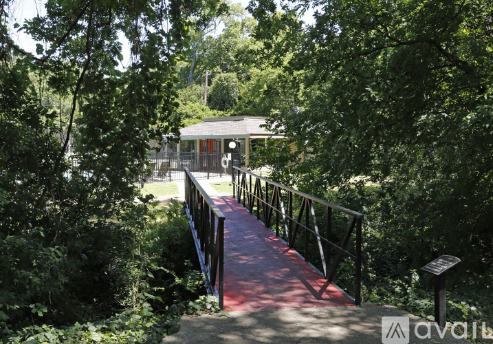 A wooden bridge with a red walkway leads to a small building in a wooded area.