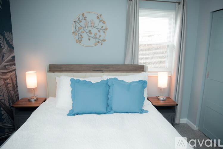 A bed with two blue pillows and a white comforter is in the foreground of a room with a decorative wall plate and two lamps on bedside tables.